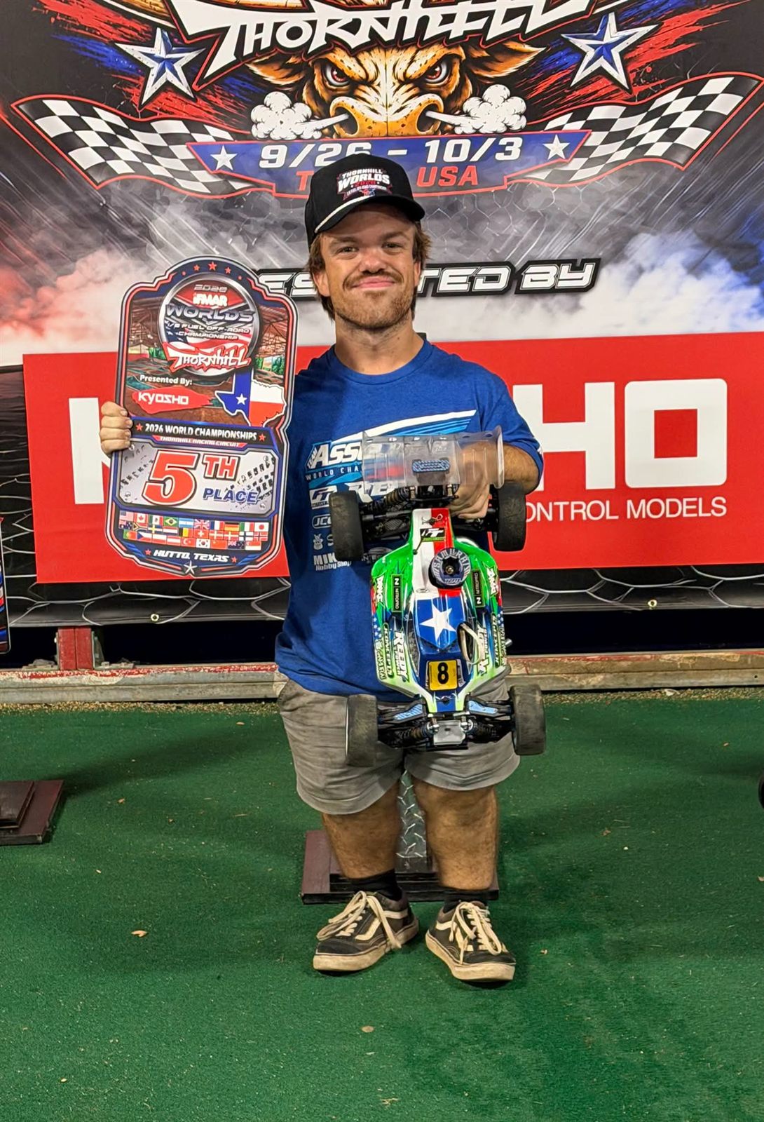 A racer holding a custom Integrity RC Designs 5th place award plaque from the 2026 IFMAR 1/8 off-road World Championship, standing with his nitro buggy in front of the event backdrop.